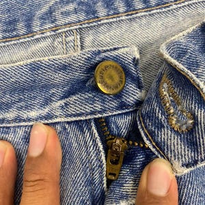 May include: Close-up of light blue denim jeans. The image shows the button, zipper, and stitching details. The button is a brass color with embossed text. The zipper is a brass color with the text "YKK" on the zipper pull.