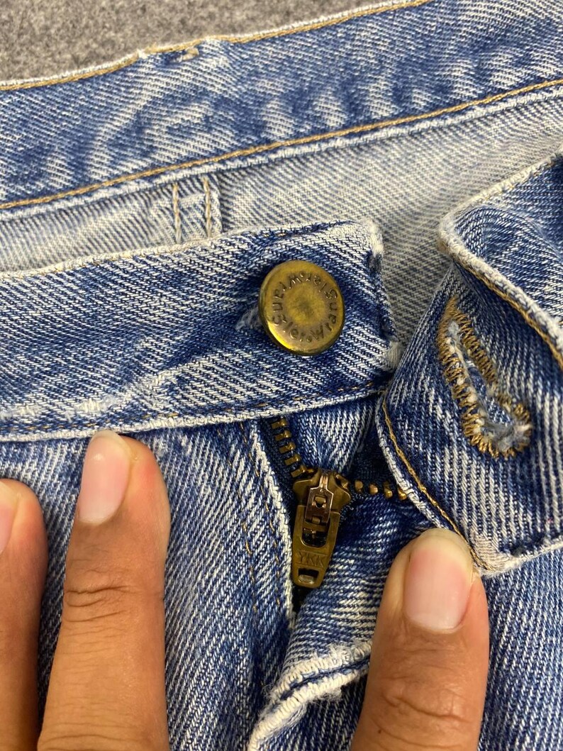 May include: Close-up of light blue denim jeans. The image shows the button, zipper, and stitching details. The button is a brass color with embossed text. The zipper is a brass color with the text "YKK" on the zipper pull.