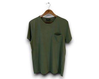 Puede incluir: Una camiseta de manga corta en color verde oliva sólido, con cuello redondo y un bolsillo de parche en el pecho izquierdo. La prenda se muestra en una percha de madera, presentando un diseño sencillo con dobladillo recto y corte estándar.