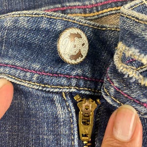 Puede incluir: Primer plano de unos vaqueros azules con cremallera de lat&oacute;n y bot&oacute;n decorativo. El bot&oacute;n tiene un dise&ntilde;o de filigrana blanca sobre fondo marr&oacute;n. Las costuras est&aacute;n acentuadas con pespuntes rosas.
