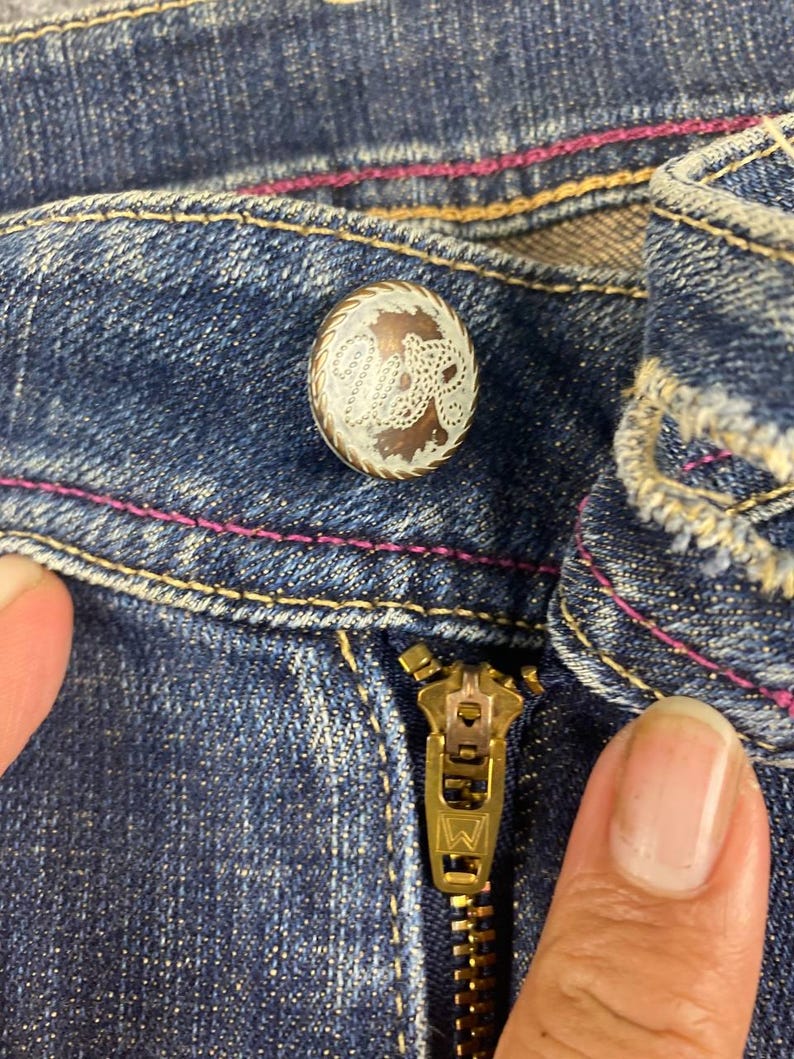 Puede incluir: Primer plano de unos vaqueros azules con cremallera de lat&oacute;n y bot&oacute;n decorativo. El bot&oacute;n tiene un dise&ntilde;o de filigrana blanca sobre fondo marr&oacute;n. Las costuras est&aacute;n acentuadas con pespuntes rosas.