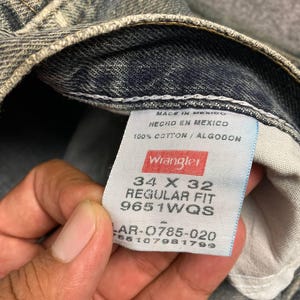 Może przedstawiać: Zbliżenie na parę wyblakłych niebieskich dżins&oacute;w. Dżinsy mają metkę z napisem "Wrangler" i zawierają rozmiar 34 x 32, regular fit i inne szczeg&oacute;ły. Dżinsy są wykonane w 100% z bawełny.