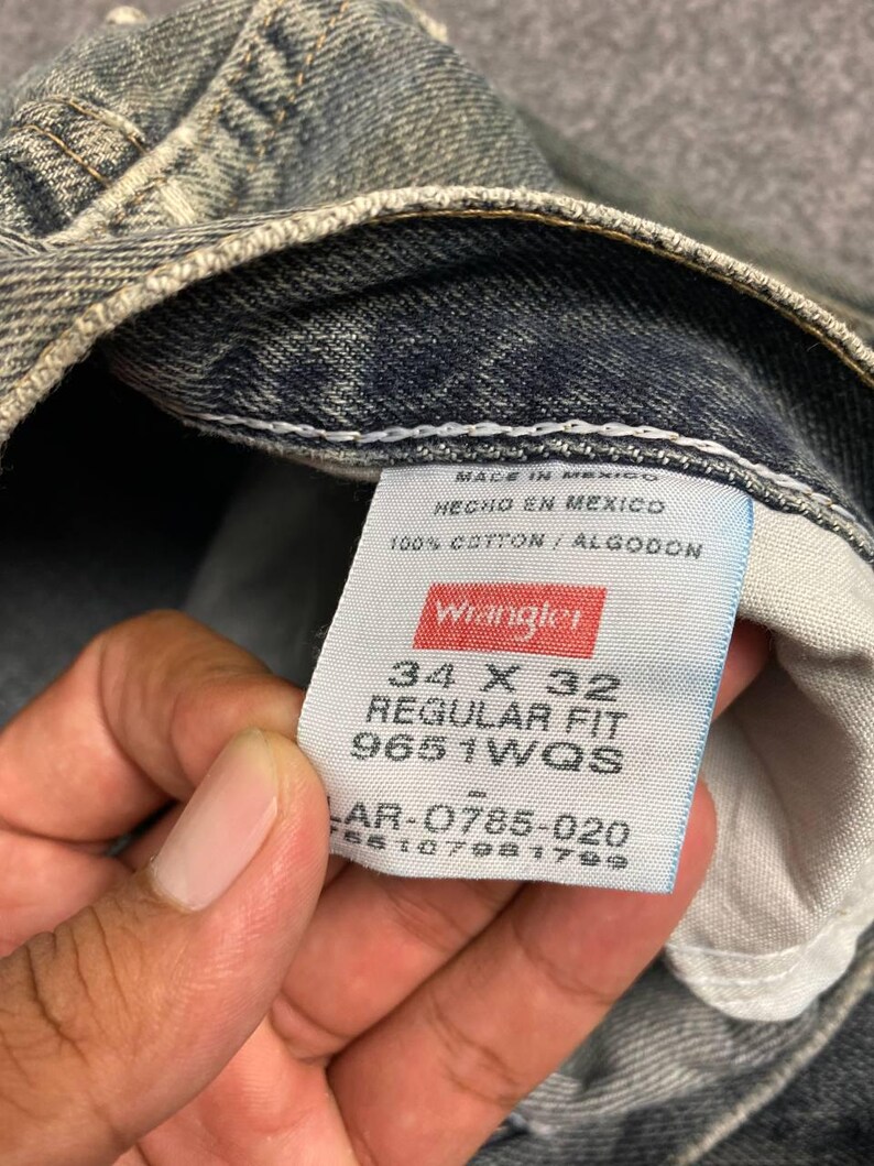 Może przedstawiać: Zbliżenie na parę wyblakłych niebieskich dżins&oacute;w. Dżinsy mają metkę z napisem "Wrangler" i zawierają rozmiar 34 x 32, regular fit i inne szczeg&oacute;ły. Dżinsy są wykonane w 100% z bawełny.