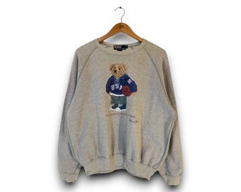 May include: A light gray Polo Ralph Lauren sweatshirt featuring a bear wearing a blue hoodie with "USA" on it, holding a basketball. The bear is wearing green pants and blue sneakers. The sweatshirt is hanging on a wooden hanger.