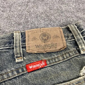 Może przedstawiać: Zbliżenie dżins&oacute;w z brązową sk&oacute;rzaną naszywką i czerwoną metką Wrangler. Brązowa naszywka ma logo i tekst Wrangler. Dżinsy są w kolorze wyblakłego niebieskiego.