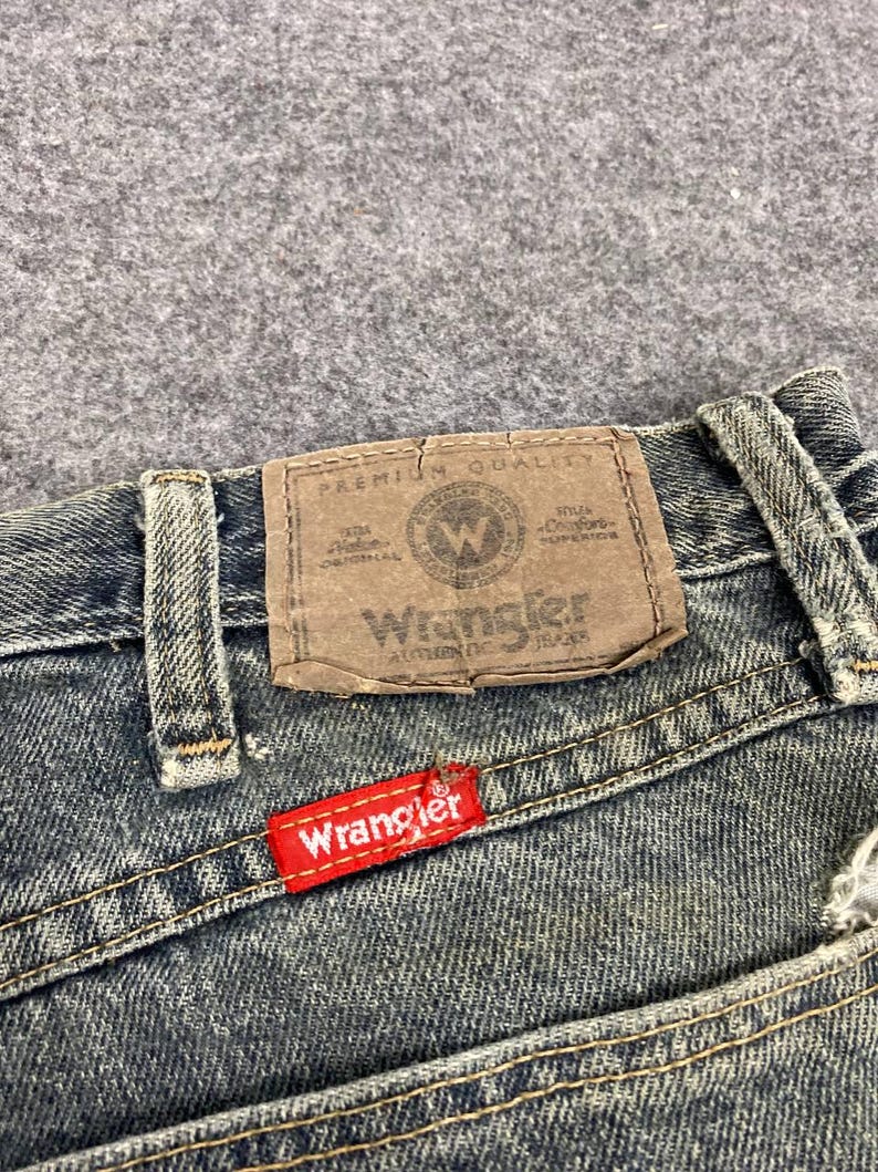 Może przedstawiać: Zbliżenie dżins&oacute;w z brązową sk&oacute;rzaną naszywką i czerwoną metką Wrangler. Brązowa naszywka ma logo i tekst Wrangler. Dżinsy są w kolorze wyblakłego niebieskiego.