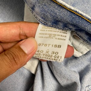 May include: Close-up of a light blue denim jeans tag. The tag reads "GREENSBORO, NC 27408 MADE IN HONDURAS OF US FABRIC HECHO EN HONDURAS CON TELA DE EE.UU. 100% COTTON/ALGODON 976T1SB 40 X 30 NS-Y602-021 68382864382.1".