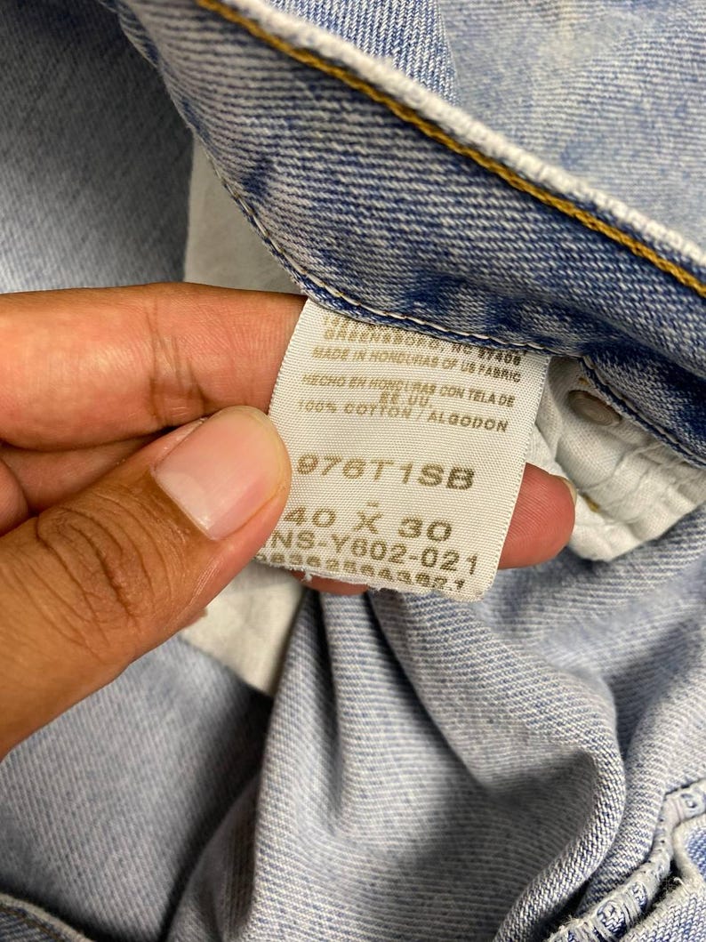 May include: Close-up of a light blue denim jeans tag. The tag reads "GREENSBORO, NC 27408 MADE IN HONDURAS OF US FABRIC HECHO EN HONDURAS CON TELA DE EE.UU. 100% COTTON/ALGODON 976T1SB 40 X 30 NS-Y602-021 68382864382.1".