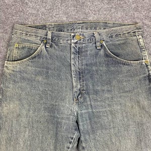 Może przedstawiać: Zbliżenie niebieskich dżins&oacute;w. Dżinsy mają zapięcie na guziki, przednie kieszenie i szlufki na pasek. Denim ma wyblakły wygląd, z jaśniejszymi obszarami wok&oacute;ł kieszeni i szw&oacute;w. Dżinsy to klasyczny, swobodny styl.
