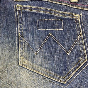 Puede incluir: Primer plano de un bolsillo de vaquero azul con costuras blancas. El bolsillo presenta un parche rectangular y un dise&ntilde;o distintivo de 'W'. Las costuras rosas acent&uacute;an la cintura.