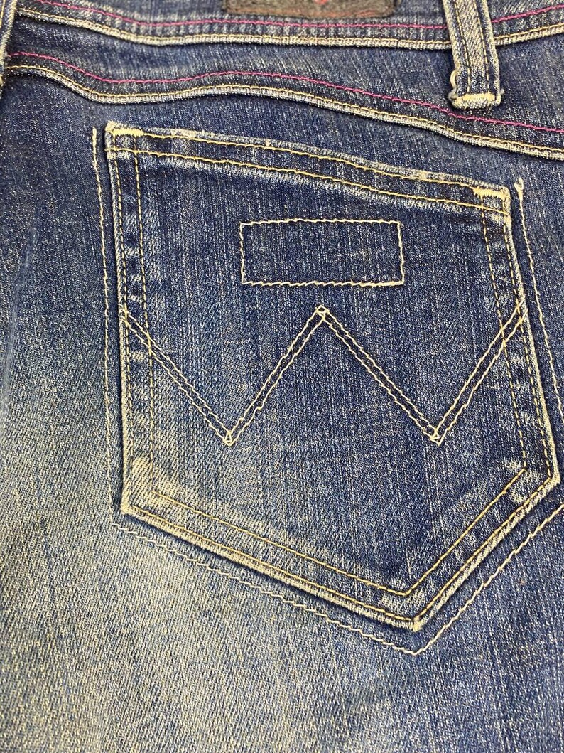 Puede incluir: Primer plano de un bolsillo de vaquero azul con costuras blancas. El bolsillo presenta un parche rectangular y un dise&ntilde;o distintivo de 'W'. Las costuras rosas acent&uacute;an la cintura.