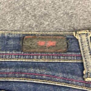 Puede incluir: Primer plano de unos vaqueros de mezclilla azules con costuras rosas y amarillas. Se ve una etiqueta rectangular gris oscuro con texto rojo. Los vaqueros tienen un dise&ntilde;o cl&aacute;sico de cinco bolsillos.