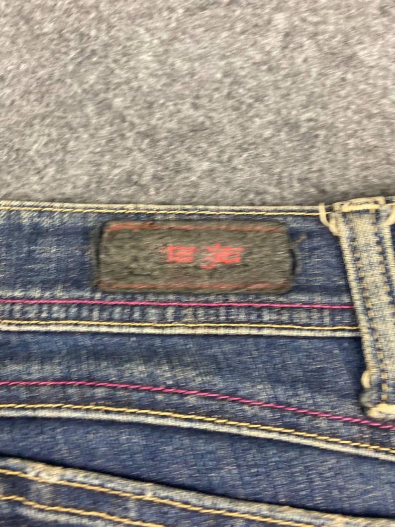 Puede incluir: Primer plano de unos vaqueros de mezclilla azules con costuras rosas y amarillas. Se ve una etiqueta rectangular gris oscuro con texto rojo. Los vaqueros tienen un dise&ntilde;o cl&aacute;sico de cinco bolsillos.