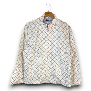May include: A long-sleeve jacket featuring a white base with a diagonal grid pattern in thin black and tan lines. The jacket has a high collar, a full-length front zipper, and is displayed on a wooden hanger against a plain white background.