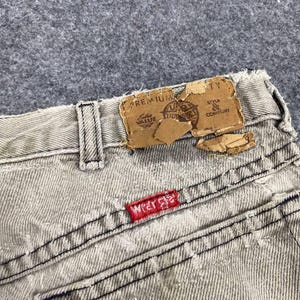 May include: Close-up of the back of light gray denim jeans. The jeans have a red "Wrangler" tag on the back pocket and a torn brown label at the waistband. The stitching is black.