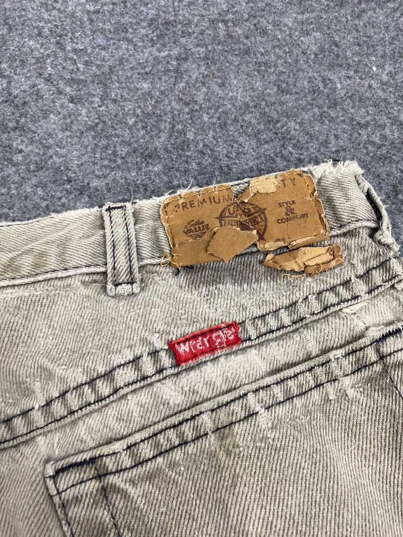 May include: Close-up of the back of light gray denim jeans. The jeans have a red "Wrangler" tag on the back pocket and a torn brown label at the waistband. The stitching is black.