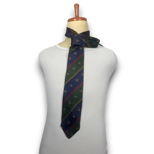 Puede incluir: Una corbata con un patrón de rayas diagonales en azul marino y verde oscuro, acentuado por finas líneas rojas y doradas. La corbata está decorada con un motivo de escudo repetitivo en toda la tela y se muestra sobre un torso de maniquí blanco.