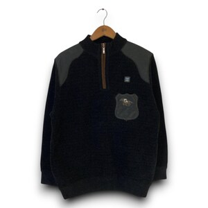 May include: A black quarter-zip pullover featuring a textured fleece-like material, dark gray shoulder panels, and a brown zipper placket. A small shield-shaped patch with an embroidered horse graphic is positioned on the left chest area.