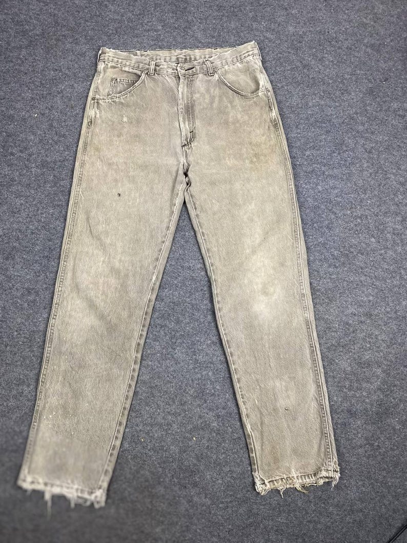 May include: Faded olive green denim jeans with a classic five-pocket design. The jeans have a straight leg cut and distressed, frayed hems. The fabric shows signs of wear and tear.