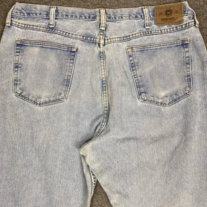 May include: Light blue denim jeans with two back pockets and a brown leather patch with the Wrangler logo. The jeans have a classic five-pocket design and a straight leg fit. The denim has a faded appearance.