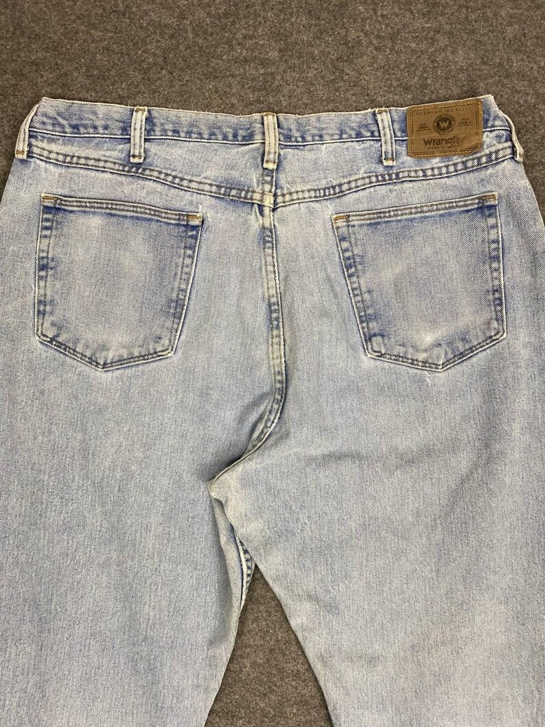 May include: Light blue denim jeans with two back pockets and a brown leather patch with the Wrangler logo. The jeans have a classic five-pocket design and a straight leg fit. The denim has a faded appearance.