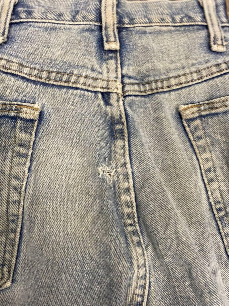 K&ouml;nnte beinhalten: Nahaufnahme der R&uuml;ckseite einer hellblauen Jeans. Das Bild zeigt den Bund, die G&uuml;rtelschlaufen und die Taschen. Der Denim hat ein verwaschenes, abgenutztes Aussehen mit einem kleinen Riss in der N&auml;he der Mittelnaht.