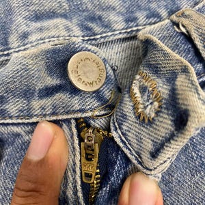 K&ouml;nnte beinhalten: Nahaufnahme von Knopf, Rei&szlig;verschluss und Denim-Stoff einer blauen Jeans. Der Knopf ist silberfarben und tr&auml;gt das Wort "Wrangler". Der Rei&szlig;verschluss ist goldfarben und tr&auml;gt die Buchstaben "YKK".