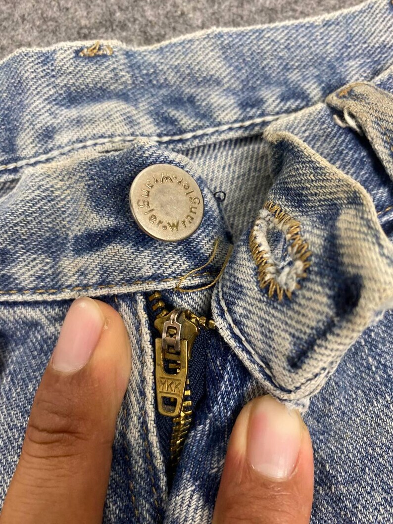 K&ouml;nnte beinhalten: Nahaufnahme von Knopf, Rei&szlig;verschluss und Denim-Stoff einer blauen Jeans. Der Knopf ist silberfarben und tr&auml;gt das Wort "Wrangler". Der Rei&szlig;verschluss ist goldfarben und tr&auml;gt die Buchstaben "YKK".