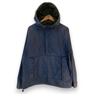 May include: A navy blue hooded anorak jacket with a front pocket and a small logo. The jacket has a drawstring at the bottom and a contrasting green lining in the hood. The jacket is hanging on a wooden hanger.
