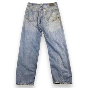 May include: Light blue denim jeans with a faded wash and distressed details. The jeans have a classic five-pocket design and a straight leg fit. The back pockets show wear and tear. The jeans are laid flat on a white surface.