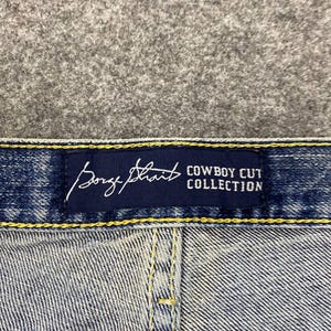 May include: Close-up of blue denim jeans with a navy blue label. The label features the brand name "Borge Strait" in a cursive font and the text "Cowboy Cut Collection" in a sans-serif font.