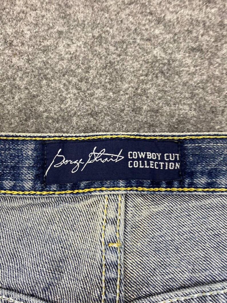 May include: Close-up of blue denim jeans with a navy blue label. The label features the brand name "Borge Strait" in a cursive font and the text "Cowboy Cut Collection" in a sans-serif font.