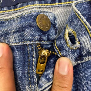 May include: Close-up of blue denim jeans with a brass zipper and button. The jeans have yellow stitching and a dark blue label that reads "Cowboy Cut Collection" and "George Strait".