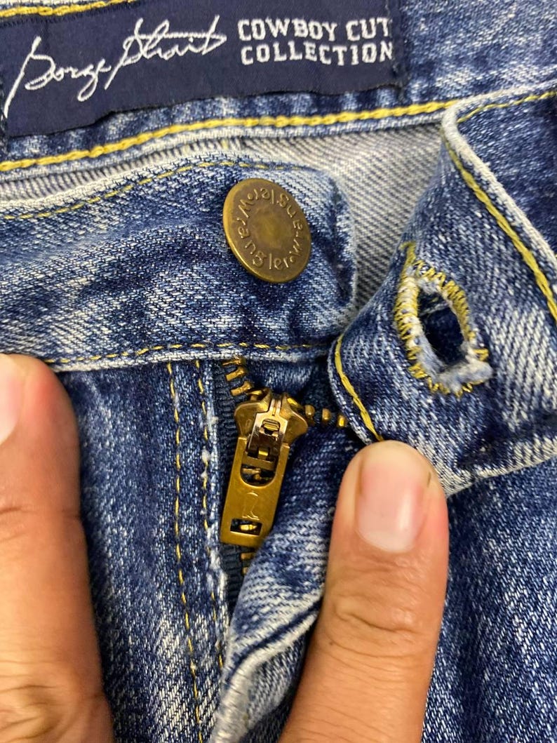 May include: Close-up of blue denim jeans with a brass zipper and button. The jeans have yellow stitching and a dark blue label that reads "Cowboy Cut Collection" and "George Strait".