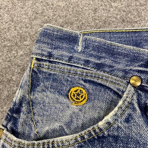 May include: Close-up of blue denim jeans with yellow stitching and a small pocket. The pocket features a yellow embroidered star design. A brass button is visible on the jeans.