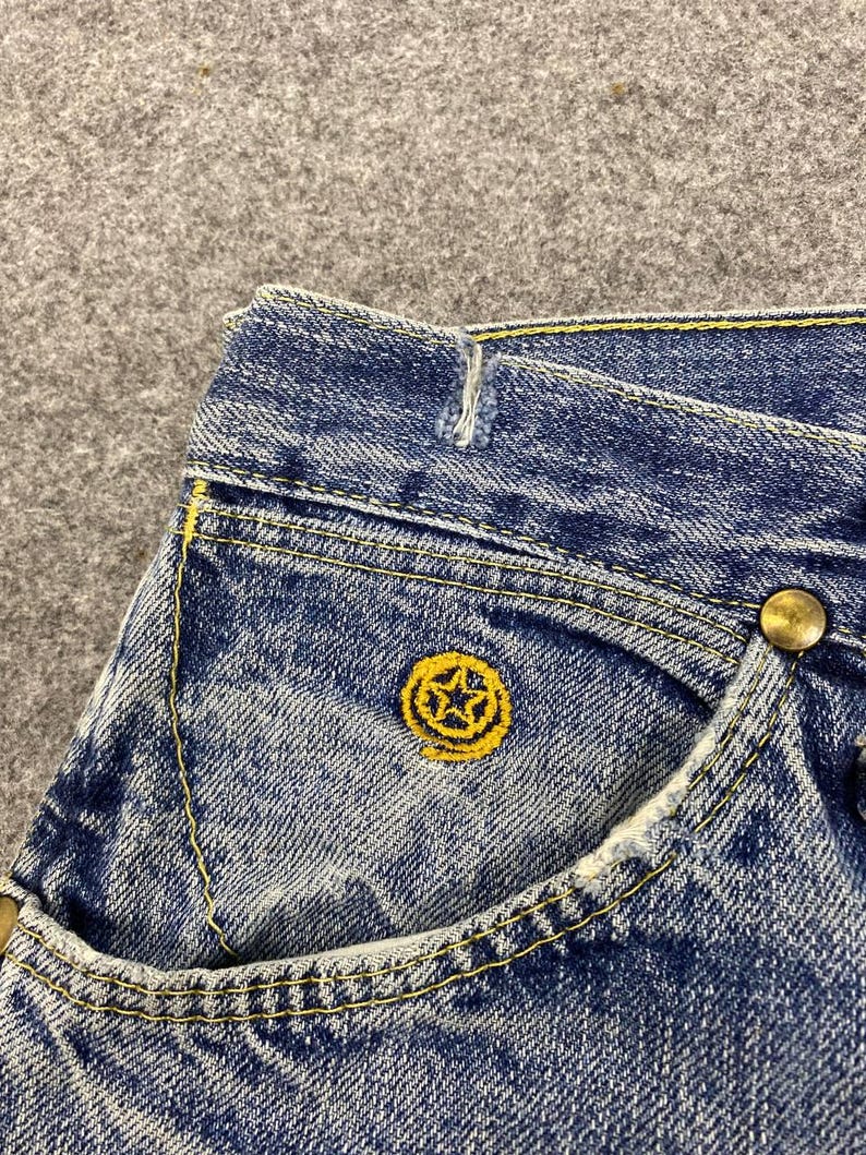 May include: Close-up of blue denim jeans with yellow stitching and a small pocket. The pocket features a yellow embroidered star design. A brass button is visible on the jeans.