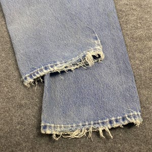May include: Close-up of light blue denim jeans with distressed, frayed hems. The jeans are folded, showcasing the texture and wear of the fabric. The frayed edges add a vintage, worn look to the garment.