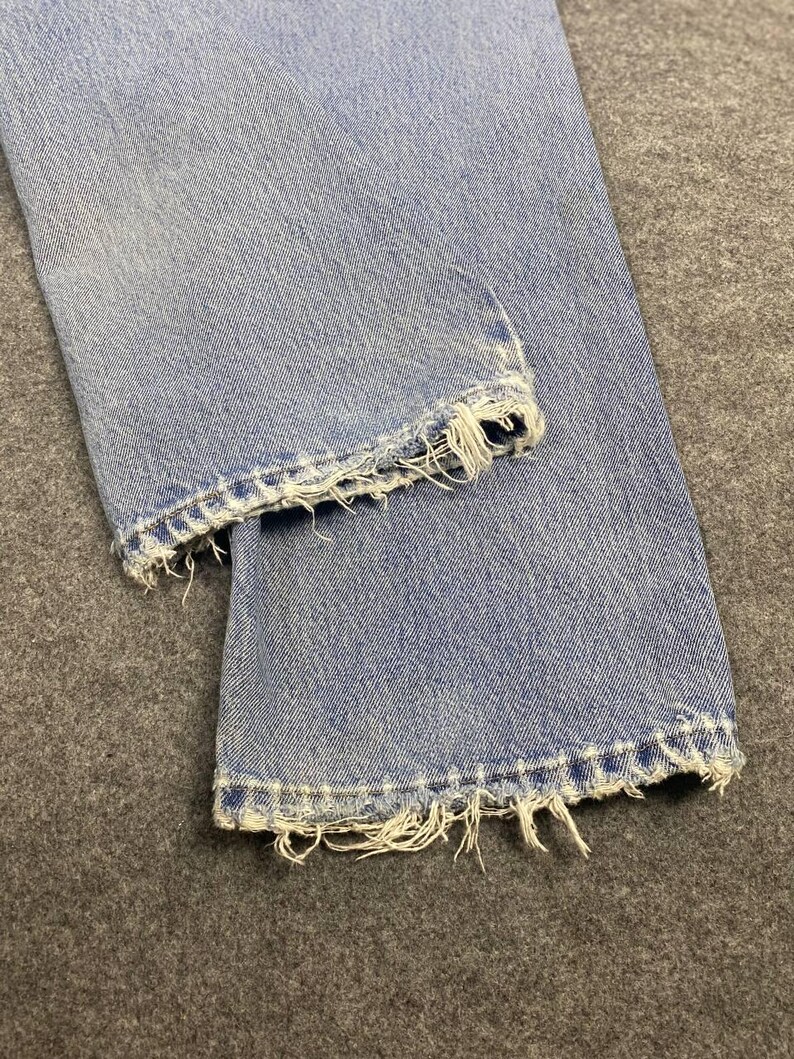 May include: Close-up of light blue denim jeans with distressed, frayed hems. The jeans are folded, showcasing the texture and wear of the fabric. The frayed edges add a vintage, worn look to the garment.