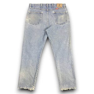 May include: Light blue denim jeans with a faded wash and distressed details. The jeans have a classic five-pocket design, a button and zipper closure, and a leather patch on the back. The jeans are a vintage style.