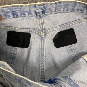 May include: Close-up of light blue denim jeans. The jeans have a classic five-pocket design with a button and zipper closure. The back pockets have black patches. The denim has a faded wash, giving it a vintage look.