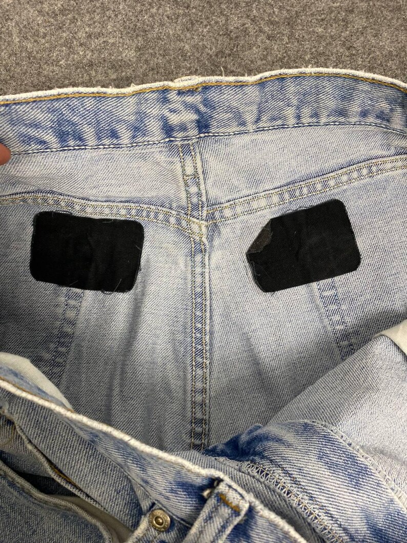 May include: Close-up of light blue denim jeans. The jeans have a classic five-pocket design with a button and zipper closure. The back pockets have black patches. The denim has a faded wash, giving it a vintage look.