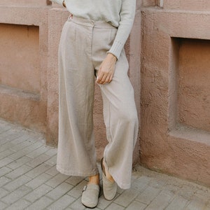 May include: A woman wearing a light gray turtleneck sweater and beige wide-leg pants with a checkered pattern. She is standing in front of a pink brick wall.