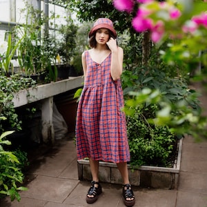 Può includere: Una donna che indossa un vestito a quadri rosso e blu con un cappello a secchiello marrone e sandali neri. È in piedi in un ambiente di serra.