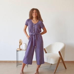 Op de afbeelding: Een vrouw draagt een paarse linnen jumpsuit met een strikceintuur en korte mouwen. Ze staat voor een witte stoel en een kleine tafel met een houten sculptuur en een witte vaas.