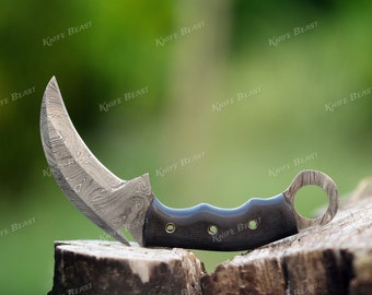 Può includere: Un coltello ricurvo color argento con un manico nero e un anello all'estremità. Il coltello è realizzato in acciaio damasco e presenta un motivo unico.