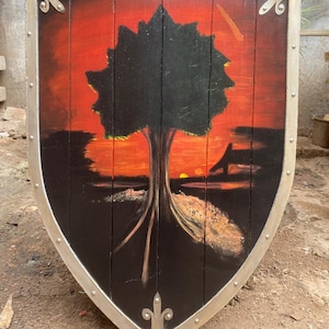 May include: A wooden shield with a pointed base, featuring a painted scene of a black tree silhouette against a vibrant orange and red sunset background. The shield is framed with a metallic silver border secured by visible rivets along the edges.