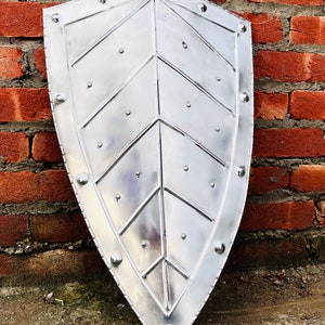 Puede incluir: Un escudo de metal plateado con un diseño en forma de hoja y remaches. El escudo está apoyado contra una pared de ladrillo.