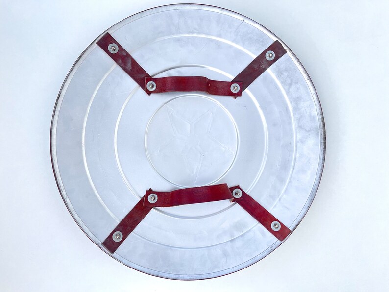 Pu&ograve; includere: Il retro di uno scudo rotondo in argento di Capitan America con cinturini in pelle rossa e rivetti metallici.
