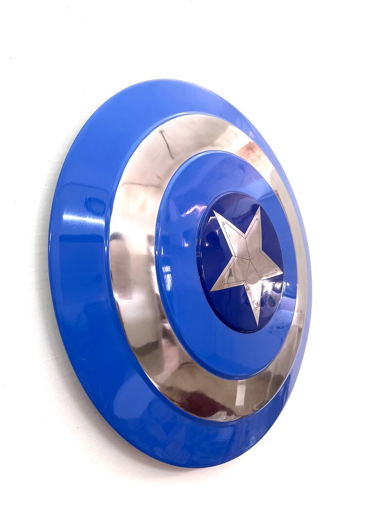Puede incluir: Un escudo azul y plateado con un dise&ntilde;o de estrella plateada central. El escudo tiene un acabado brillante y una forma circular, con anillos conc&eacute;ntricos azules y plateados. Es un accesorio o art&iacute;culo de colecci&oacute;n.