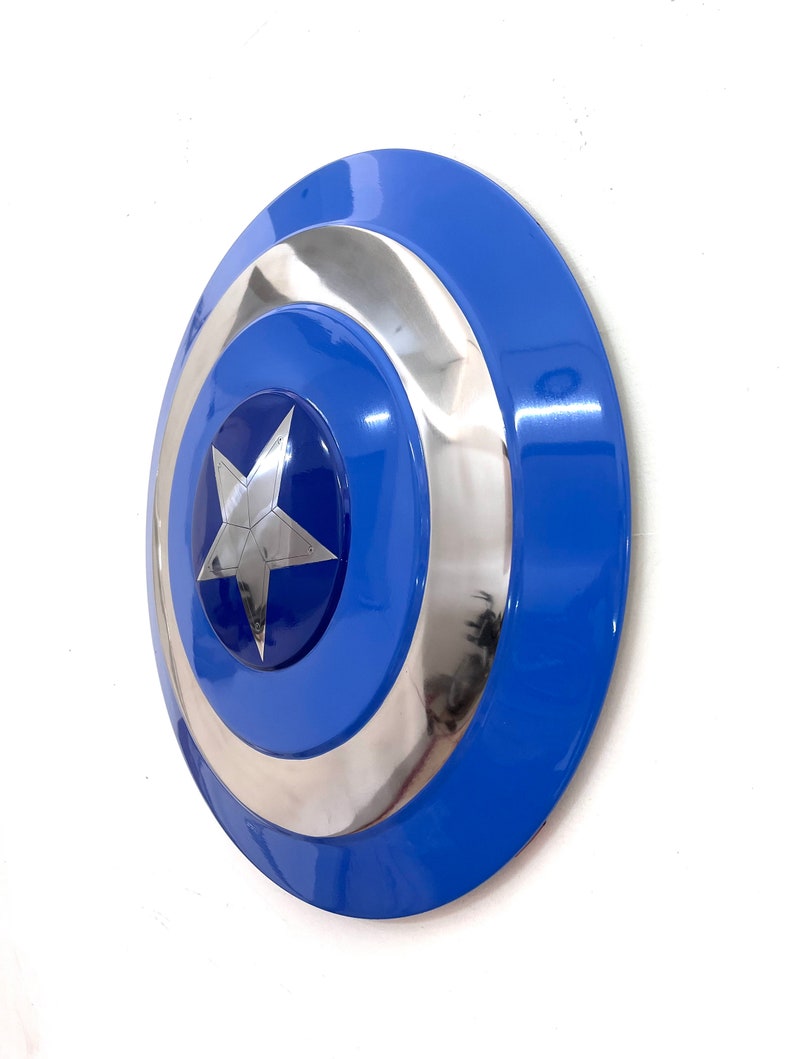Puede incluir: Un escudo azul y plateado con un emblema de estrella. El escudo tiene un dise&ntilde;o circular con un anillo exterior azul, un anillo central plateado y un centro azul con una estrella plateada. El escudo es un accesorio.